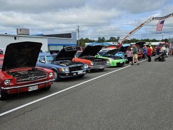 Classic cars lined up at outdoor car show with people walking and fire truck in background holding American flag